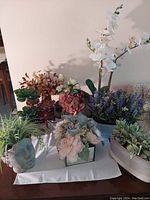 Photo showing a variety of artificial flower arrangements including orchids, succulents, and mixed floral bouquets in different decorative pots and containers on a white surface against a plain wall.