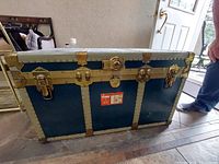 Front view of the vintage metal travel trunk showing blue panels, silver reinforcements, and gold-toned hardware.