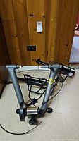 CycleOps gray indoor bike trainer frame showing sturdy base, resistance roller, and adjustment cable on floor against wood paneling.