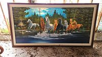 Full view of the large framed painting showing seven horses running through water with trees in the background, resting on the ground.