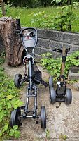 Two golf caddies outdoors on concrete surrounded by plants and tree stump. One caddy is black and gold with three wheels, the other is black with four wheels and a mesh bag attached.