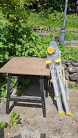 Worktable and folded extension ladder shown outdoors on concrete surface with green shrubbery behind.