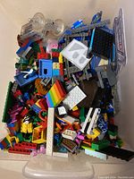 Photo showing a bin filled with assorted LEGO pieces in various colors and shapes including bricks, plates, vehicle parts, stickers, and small accessories.