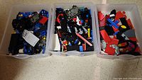 Three plastic bins filled with assorted loose Lego pieces in various colors and shapes, placed on a floor surface.