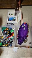 Three bins with assorted Lego pieces and a large purple Lego boat hull. One bin contains mixed bricks and plates, another contains various Lego components including wheels and a minifigure.