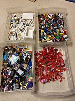 Four plastic bins containing assorted LEGO bricks sorted by colors: white and light colors, multi-color (including purple and black), red pieces, and assembled vehicle parts.