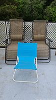 Photo showing two brown woven fabric lounge chairs with metal frames and armrests side by side with a small blue fabric beach chair in front on concrete surface outdoors.