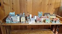 Wide view of 17 miniature buildings arranged on a wooden shelf, showing different architectural styles and sizes.