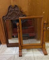 Two wooden framed mirrors side by side, one large wall mirror with carved top and one smaller tabletop mirror with tilt supports