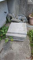 Stack of eight square concrete paving stones outdoors, stacked on ground near wall with some greenery around.