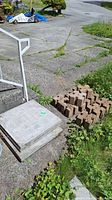 Four concrete paving stones stacked on top of each other on a cemented outdoor area. Adjacent to the stones is a group of concrete bricks arranged neatly on the ground.