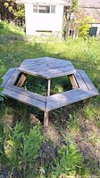 Photo showing wooden hexagonal picnic table placed outdoors on grassy ground, weathered surfaces visible.
