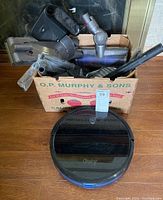 Eufy RoboVac 115 robotic vacuum placed on wooden floor alongside a cardboard box containing various vacuum cleaner attachments and nozzles.