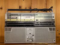 Front view of Sharp GF-555 dual cassette stereo recorder with visible dual cassette decks labeled "Tape 1" and "Tape 2", speaker grills, handle, and control panel with buttons and knobs.
