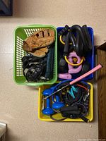 Top view of three plastic baskets containing a child's baseball mitt, black protective headgear, hand weights, straps, a pink bat and other items.
