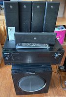 Front view of the Samsung AV receiver with four tall black surround speakers on top and a remote in front of the receiver. Subwoofer positioned below the receiver.