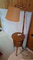 Wooden lamp table with round top, fabric lampshade, and turned wooden stand. Shows stains and signs of wear. Lamp cord visible on floor.