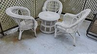 Three white wicker chairs and one round wicker side table set on concrete floor outside with lattice fence behind.