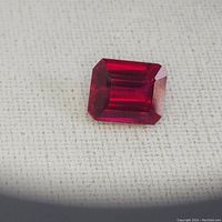 Close-up image of emerald cut red ruby gemstone on a white textured background, showing facets and color.