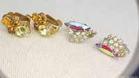Two pairs of vintage clip earrings: yellow Czech rhinestones and aurora borealis crystals with clear rhinestones on a white fabric background.