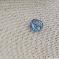 Photo of a single loose blue aquamarine gemstone on beige fabric background, showing oval facets and light blue color.