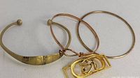 Close-up photo showing three items: engraved open cuff, a thin copper-colored bangle, and a thin gold-colored bangle with an Egyptian Eye of Horus pendant on the copper bangle.