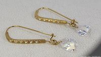 Close-up photo showing gold-tone rectangular hoop earring with embedded clear rhinestones and a single clear pear-shaped dangling crystal.