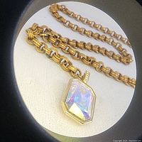 Close-up photo showing gold-tone chain and large iridescent faceted rectangular pendant with safety lock bail.