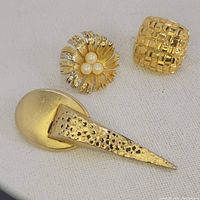 Three gold-tone single clip earrings in varied designs displayed on white fabric background.
