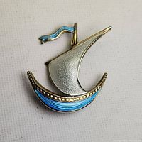 Front view of boat-shaped brooch showing silver sail, blue enamel hull with beading, and blue enamel flag.
