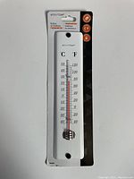 Front of the thermometer in original packaging, showing white plastic rectangular body with glass/plastic thermometer tube and dual Celsius/Fahrenheit scale markings.