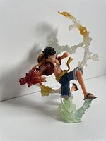 Front and side views of Monkey D Luffy figure with red flame effects on right arm, wearing open red shirt, blue shorts, straw hat on back, and sandals. Translucent flame and smoke effects swirl around the figure with yellow and green hues on the base.