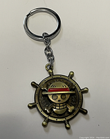 Front side of the metal One Piece keychain showing the straw hat skull logo on a ship's steering wheel design.