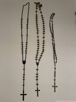 Three traditional rosary necklaces with black beads and cross pendants laid out on a white surface. The crosses are black and silver in color.