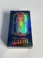 Front view of the sealed A. E. Waite Tarot cards deck box with holographic illustration showing a robed figure and the letters B and J.