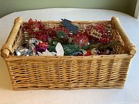 Wicker basket filled with assorted Christmas craft supplies, showing greenery, floral, and decorative elements.