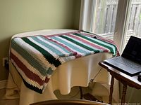 Crocheted blanket draped on a flat surface showing its striped pattern and size relative to a laptop and table.