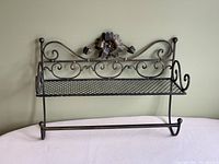 Full view of black metal bathroom shelf showing mesh bottom, scroll design on back and sides, flower decoration at top center, and towel bar below.