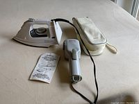 Proctor Silex steam iron, Sunbeam Steam Valet garment steamer with cord, accompanied by carry case and instruction manual on beige surface.