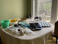 Overview of multiple kitchen items on table including bowls, strainers, kettle, coffee pot, muffin trays, loaf pan, butter dishes, and some measuring cups and spoons.