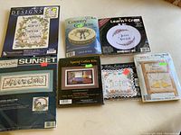 Top view of all 7 cross stitch kits and 1 mounting board arranged on a table, showing brand and design pictures on kit packaging