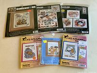 Full view of six packaged cross stitch kits from brands Dimensions and Janlynn showing various cat and floral designs.