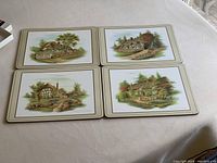 Four placemats laid out, each showing a different painted scene of English country cottages with garden settings.