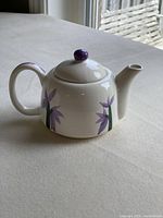 Single cup white ceramic teapot with purple flower design and purple knob on lid, shown closed on table.