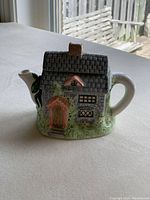 Front and side view of single cup teapot shaped like a house with painted door, windows, and greenery detail