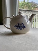 Side view of white ceramic teapot showing blue rose floral design with handle and spout visible