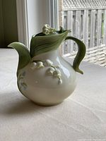 Side view of teapot showing the white ceramic body with green sculpted leaves on handle and spout, and white Lily of the Valley flowers on the surface.