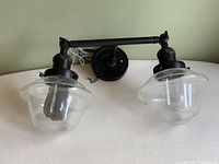 Front view of the two-light vanity lamp showing the black metal frame and glass shades, the bulbs within the glass shades are visible.
