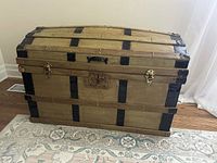 Front view of the wooden trunk showing the curved top, metal bands, clasps, and overall rectangular shape.