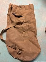 Full view of the brown army canvas duffle bag laid flat on a surface, showing the shoulder strap, metal buckle and general shape.